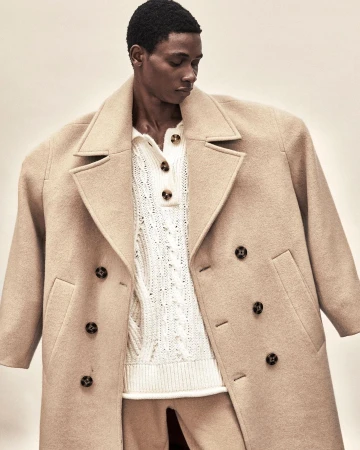 Black man wearing an oversized camel wool coat and white cable knit sweater against a plain background