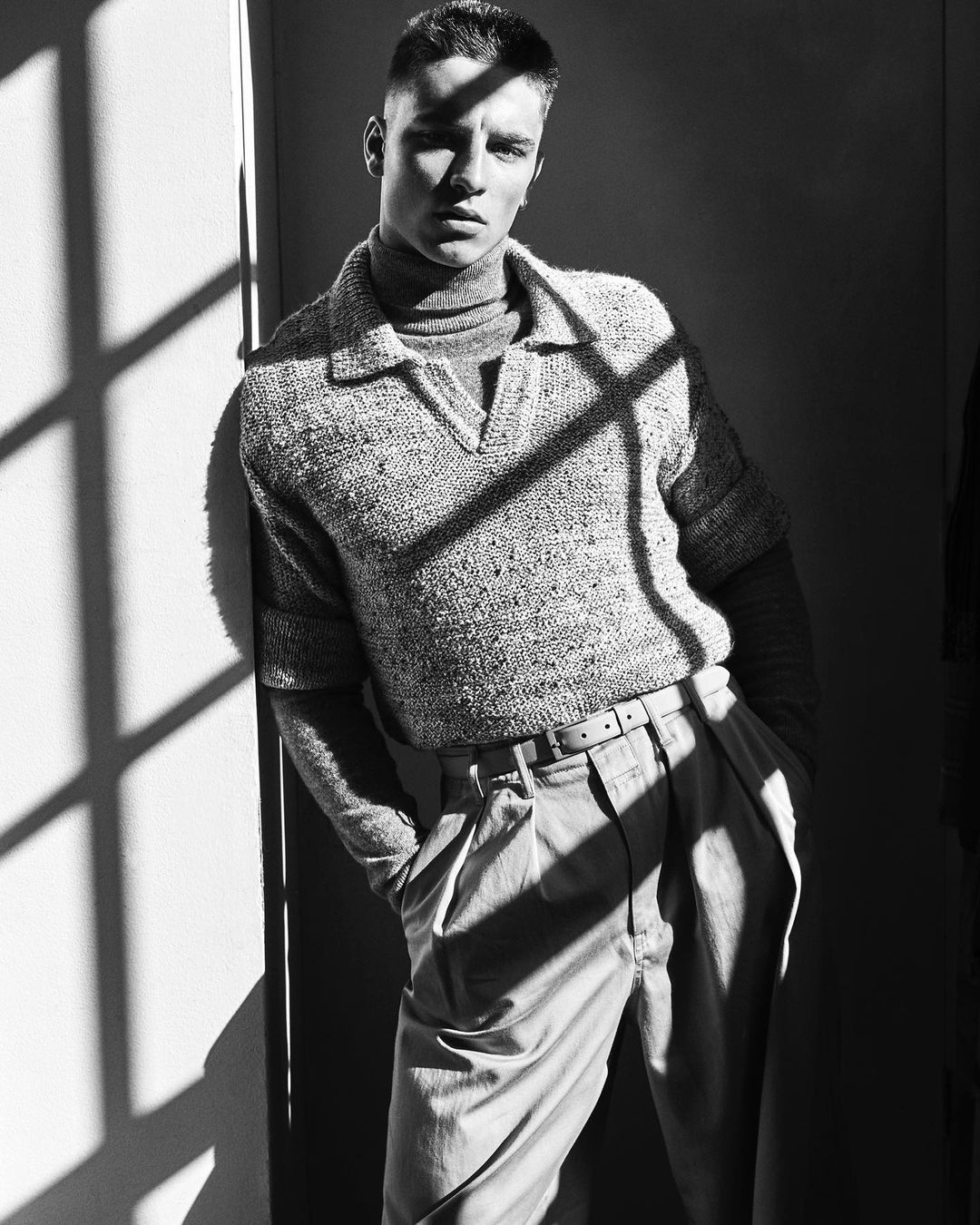 Monochrome portrait of a young man leaning against a wall in neutral-toned casual wear, with sunlight creating shadows.