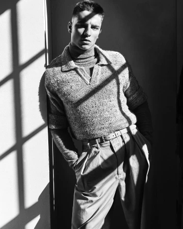 Monochrome portrait of a young man leaning against a wall in neutral-toned casual wear, with sunlight creating shadows.