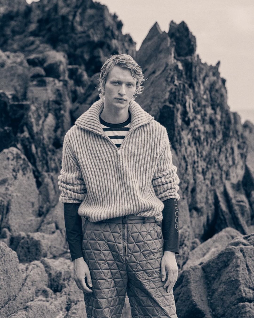 Male fashion model stands on rocky beach in a ribbed zip-up sweater and quilted pants, posing in front of large rock formations.