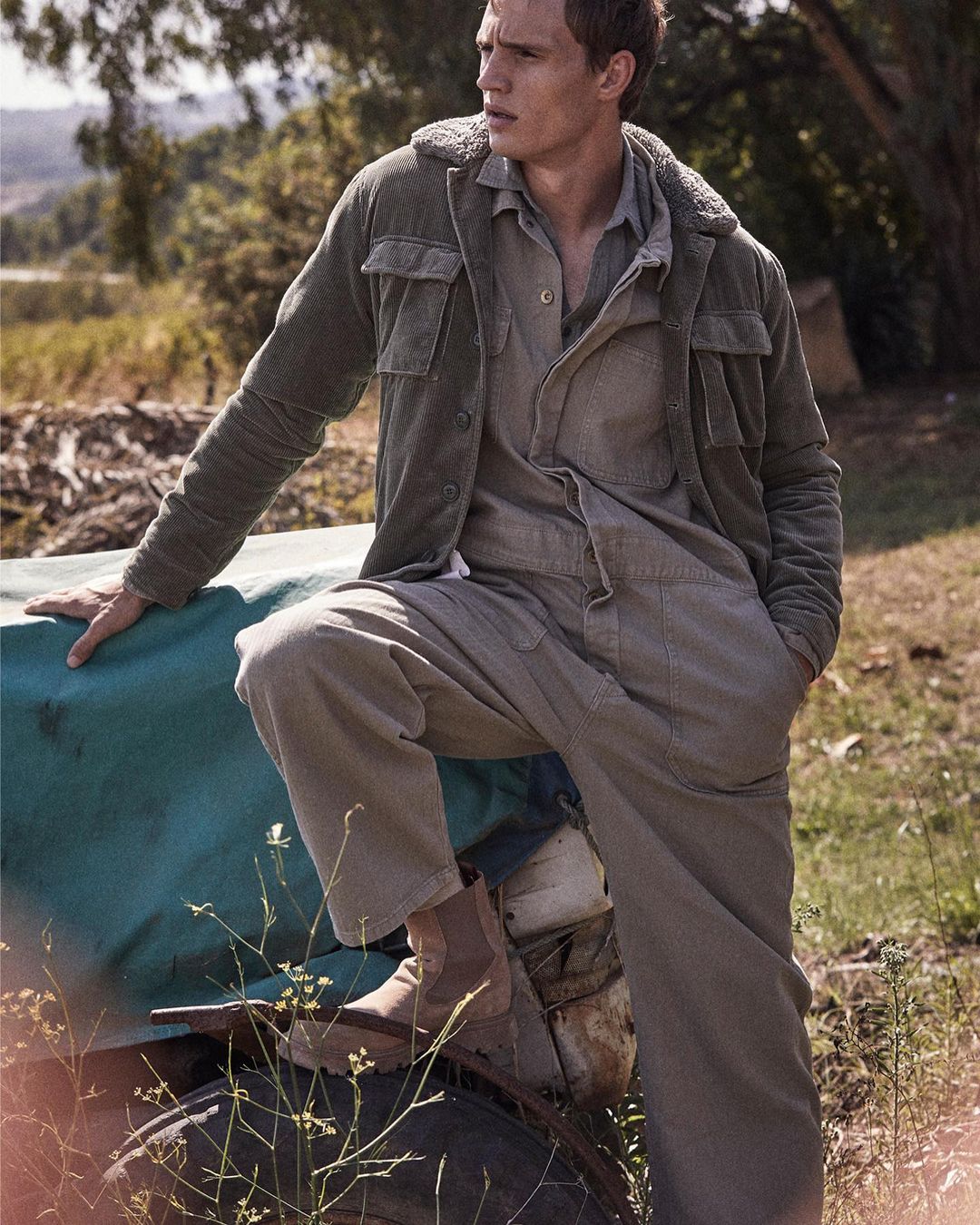 Man in workwear-inspired fashion posing outdoors, wearing a jacket, jumpsuit, and boots, with a rural background featuring a field.