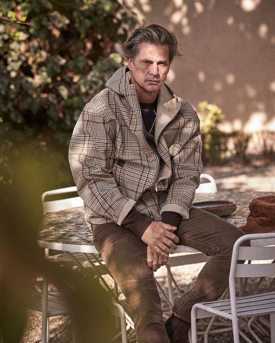 Mature adult male sits outdoors at a white table with neutral background. He wears a patterned jacket, brown pants, and black shoes.