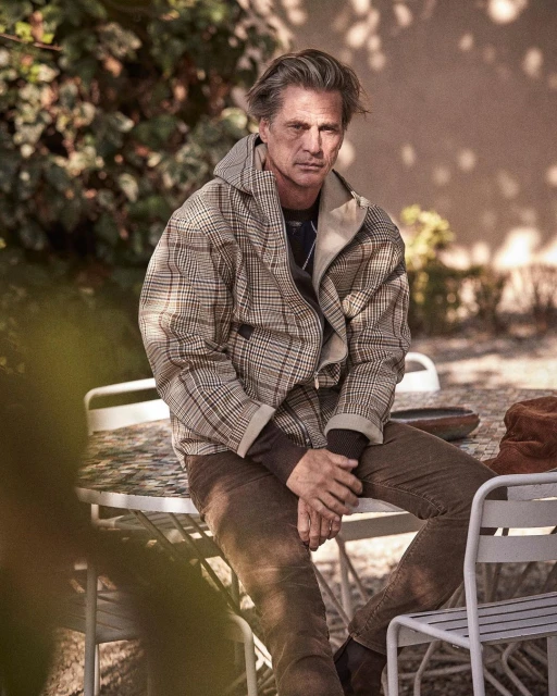 Mature adult male sits outdoors at a white table with neutral background. He wears a patterned jacket, brown pants, and black shoes.