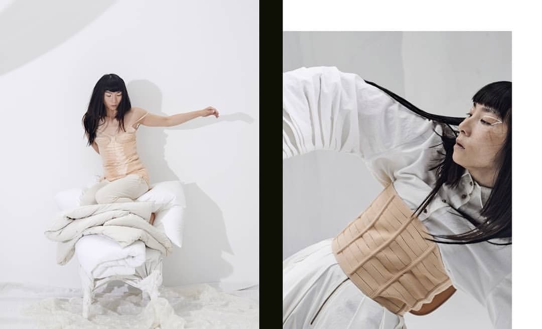 An Asian woman with dark hair poses in a studio fashion shot, perched on a chair covered in cushions, wearing a light-colored slip dress.