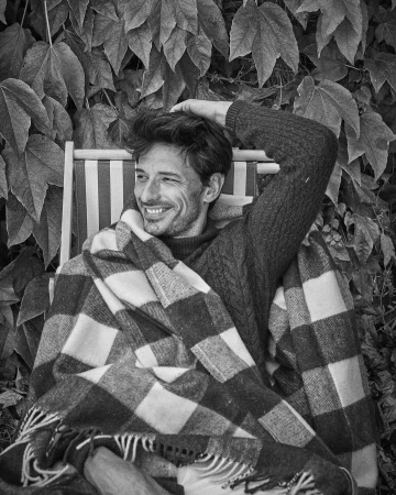 Monochrome portrait of a smiling man with hand behind head, sitting in a chair with a blanket, backdrop of dense leafy foliage.
