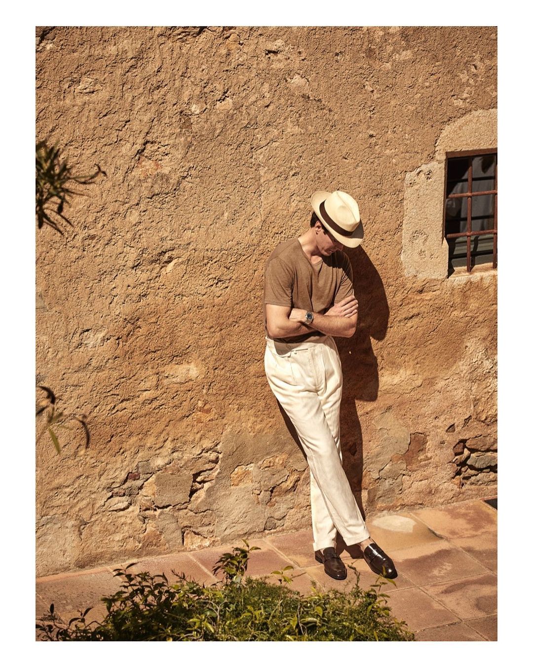 Man in a hat leans against a textured wall outdoors, wearing cream pants and a brown shirt, his arms crossed with a wristwatch.