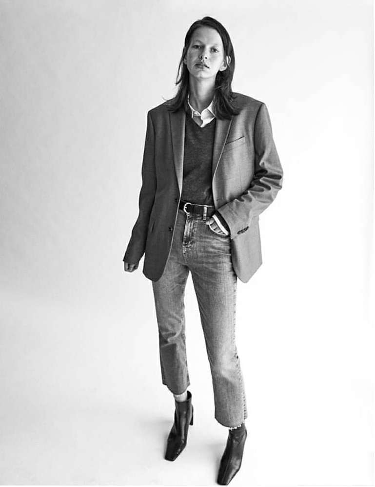 A full shot of a woman in a blazer, jeans, and boots against a white background. Fashion editorial photography in black and white.
