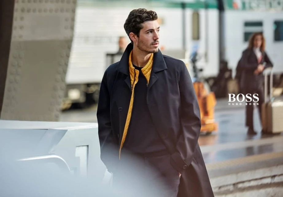 A stylish man in a black coat and mustard vest stands near train tracks on a platform, with a blurred train in the backdrop and BOSS logo.