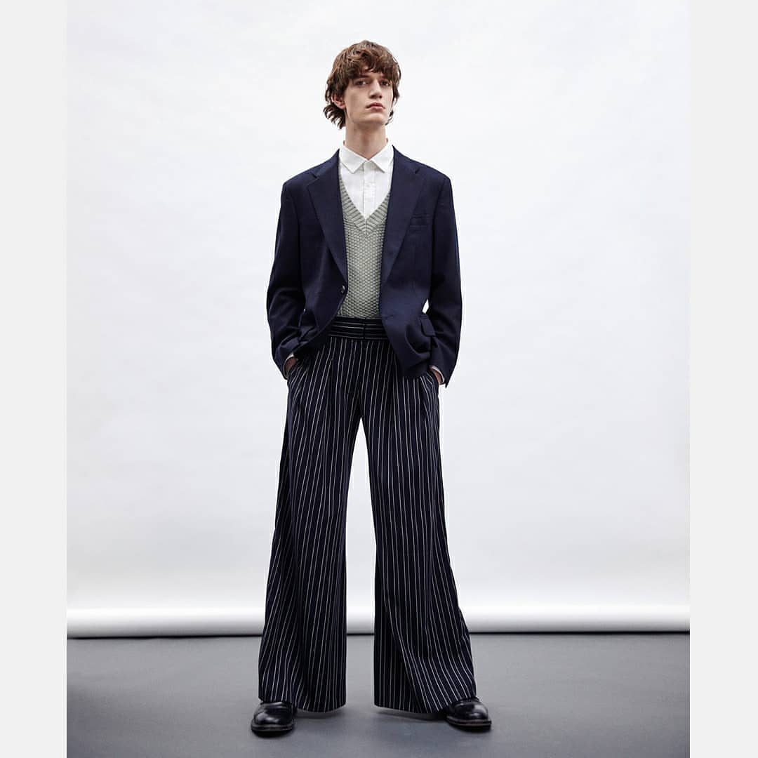 Full shot of a light-skinned male model posing in a navy blazer, button-down shirt, knit vest, and wide-legged, vertically striped trousers.