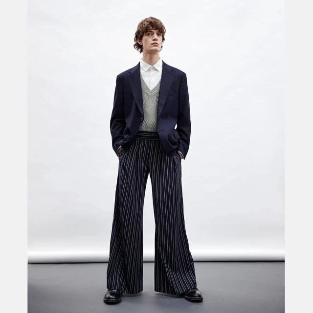 Full shot of a light-skinned male model posing in a navy blazer, button-down shirt, knit vest, and wide-legged, vertically striped trousers.
