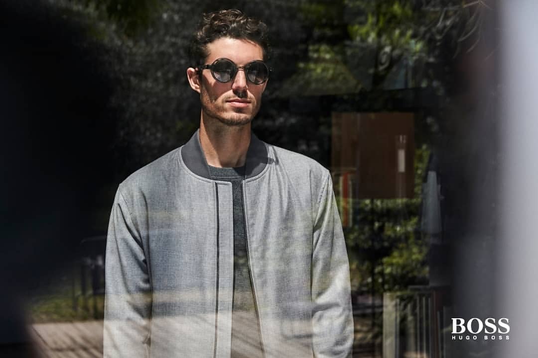 Portrait of a stylish adult male with sunglasses wearing a grey jacket standing outdoors with natural lighting and a blurred background.
