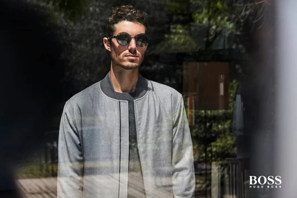 Portrait of a stylish adult male with sunglasses wearing a grey jacket standing outdoors with natural lighting and a blurred background.
