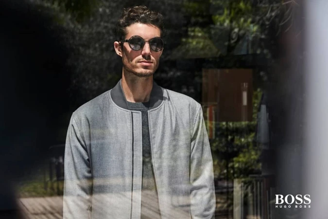 Portrait of a stylish adult male with sunglasses wearing a grey jacket standing outdoors with natural lighting and a blurred background.
