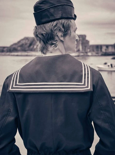 Back view of an adult male wearing a dark sailor uniform and cap looking out towards the water, with rocky land in the background.