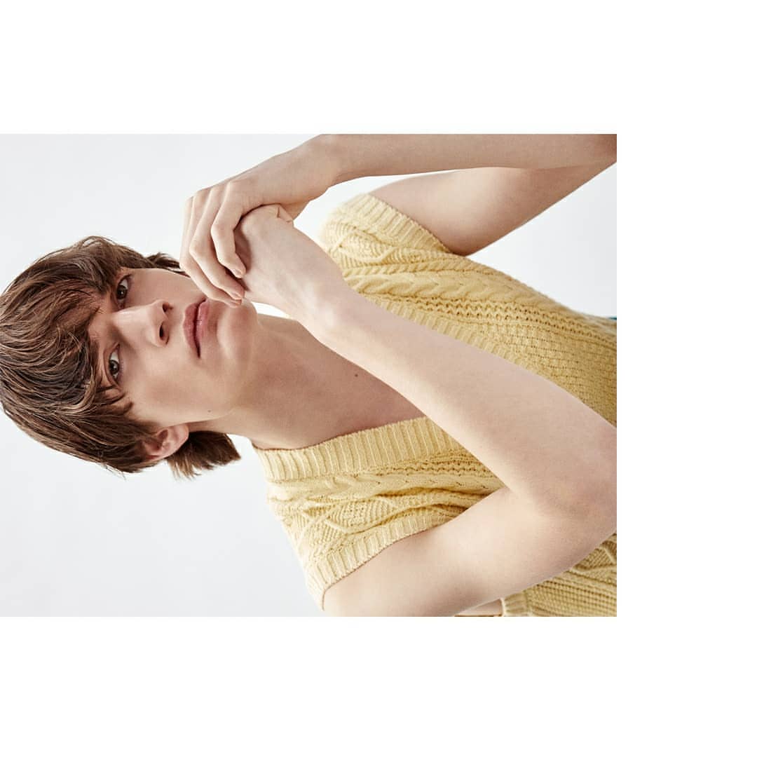 Portrait of a young adult male with brown hair, wearing a yellow knit shirt, posing against a bright white background in a fashion shot.