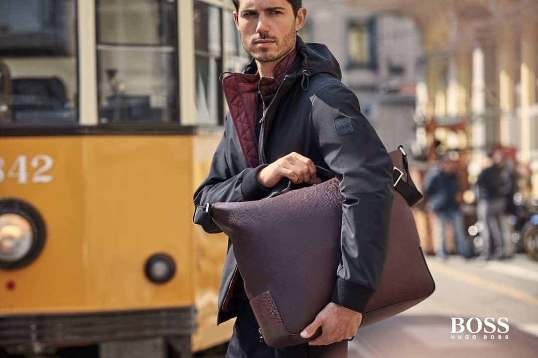 Male fashion model in a hooded jacket carries a leather bag in an outdoor urban setting with a yellow tram in the background.
