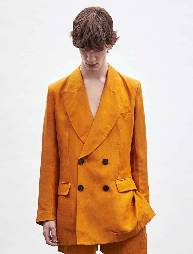 Male model in orange double-breasted suit against white backdrop in a medium shot. Fashion forward, neutral expression, studio lighting.