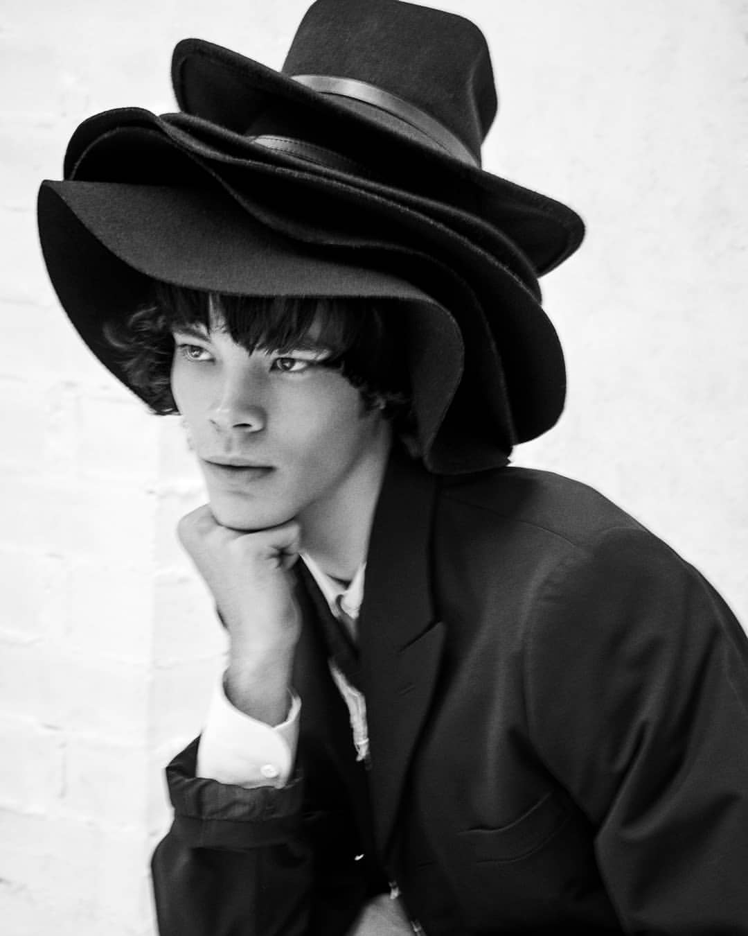 Monochrome portrait of a person wearing an oversized stacked hat with a black blazer, posing in front of a white brick wall.