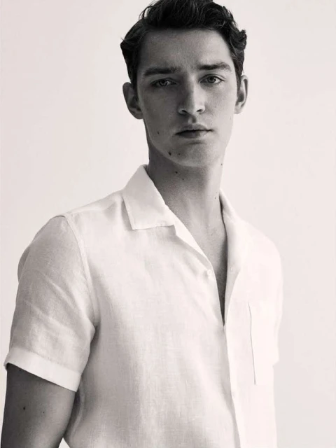Monochrome portrait of a young adult male with dark hair, wearing a white linen shirt, looking directly at the camera, studio lit.