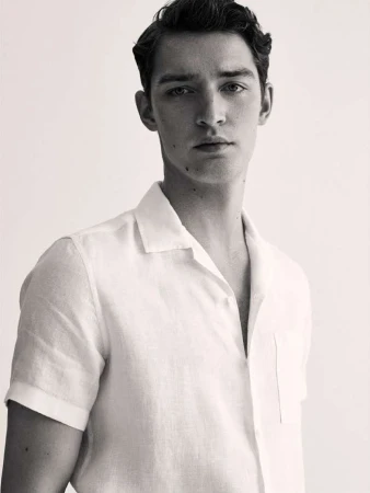 Monochrome portrait of a young adult male with dark hair, wearing a white linen shirt, looking directly at the camera, studio lit.