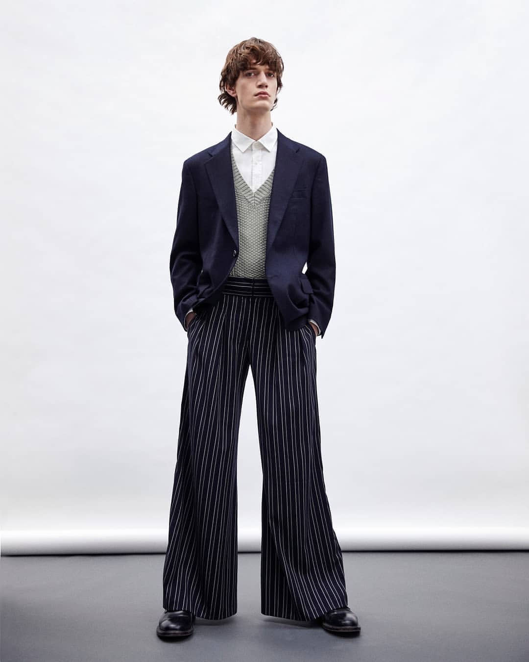 Male fashion model in studio wearing navy blazer, knit vest, white shirt and striped wide-leg pants, hands in pockets.