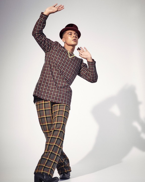 Stylish individual in a plaid suit and hat strikes a dynamic pose in a photo studio, showcasing a blend of vintage and contemporary fashion.