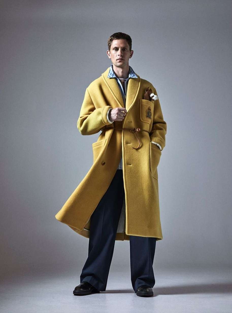 A stylish man wearing a long yellow coat and wide navy pants poses in a studio with soft lighting, showcasing a blend of classic and modern fashion.
