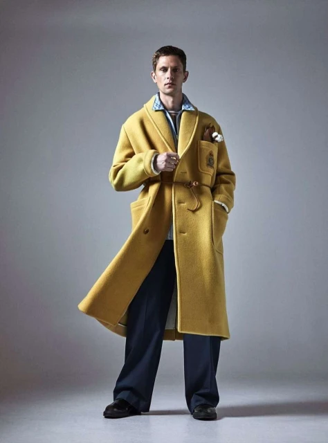 A stylish man wearing a long yellow coat and wide navy pants poses in a studio with soft lighting, showcasing a blend of classic and modern fashion.