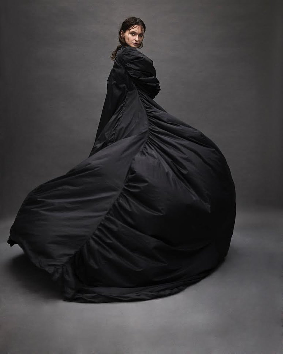 Fashionable woman posing in voluminous black coat in studio, looking over her shoulder with a neutral expression, on grey background.