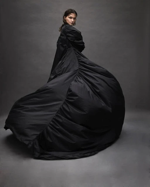 Fashionable woman posing in voluminous black coat in studio, looking over her shoulder with a neutral expression, on grey background.