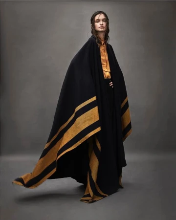 Fashionable adult woman in a mustard-yellow top and black cape with tan stripes poses in a photo studio with soft neutral lighting.