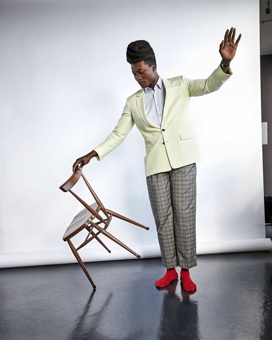 Stylish Black male balances chair in studio, wearing blazer, patterned slacks, red socks, creating a dynamic and fashionable pose.