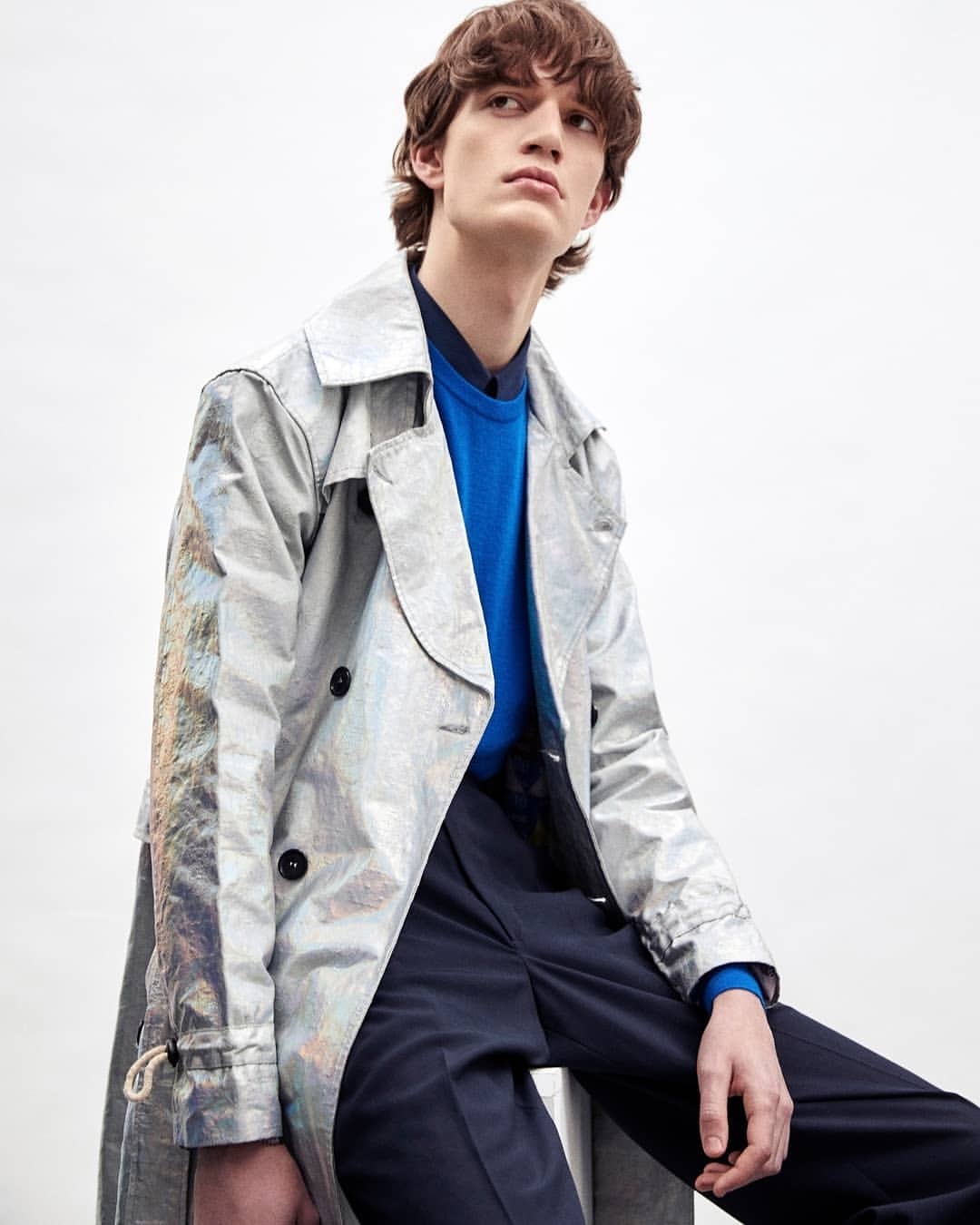 Fashion portrait of a young adult male with wavy brown hair wearing a silver trench coat and a blue sweater against a white background.