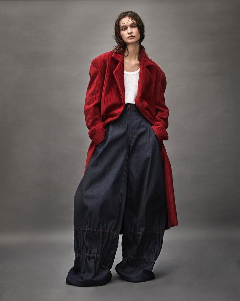 Model wearing a red coat and wide dark trousers standing against a seamless grey backdrop photographed by Diego Merino