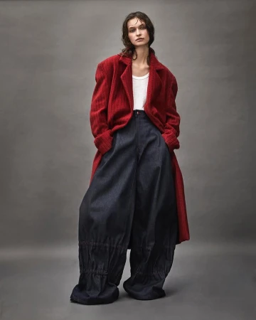 Model wearing a red coat and wide dark trousers standing against a seamless grey backdrop photographed by Diego Merino