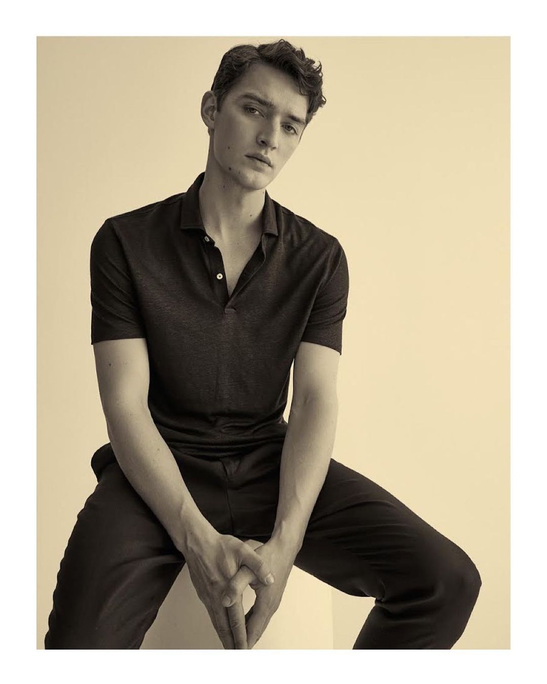 Portrait of a young adult man in a polo shirt and dress pants, seated indoors, with a neutral facial expression in sepia tones.