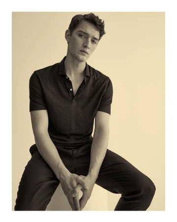 Portrait of a young adult man in a polo shirt and dress pants, seated indoors, with a neutral facial expression in sepia tones.
