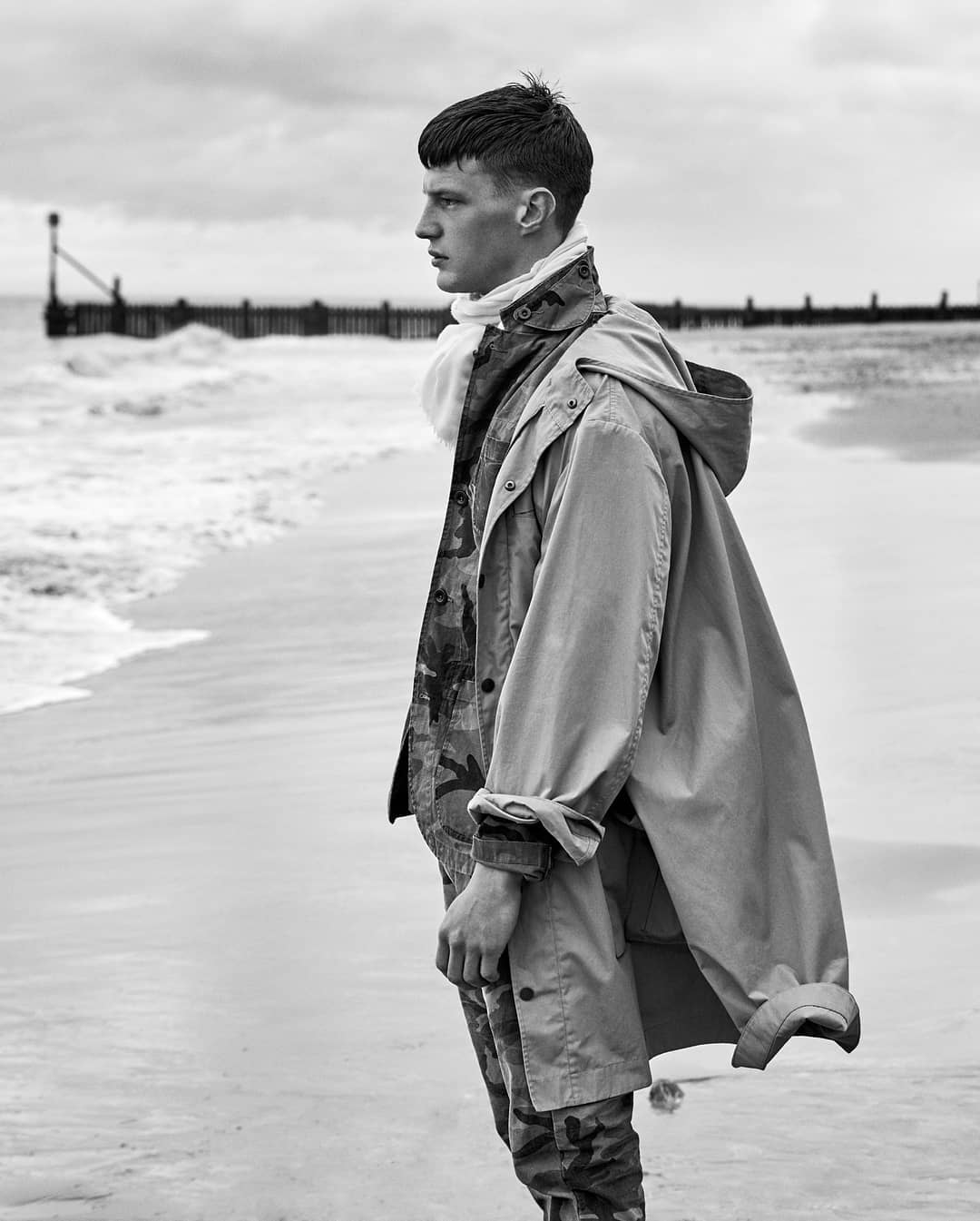 Black and white portrait of a young adult male standing on a beach in a hooded jacket and camouflage pants, looking out towards the ocean.