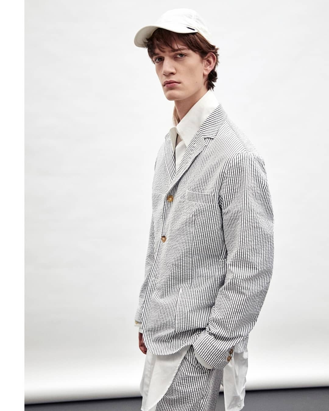 A young adult male poses in a photo studio wearing a white baseball cap, a white dress shirt, and a pinstripe blazer and pants suit.