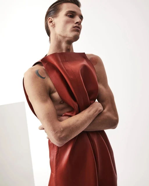 Portrait of a muscular adult male model with fair skin and slicked-back hair, wearing a rust-colored leather garment in a white studio.