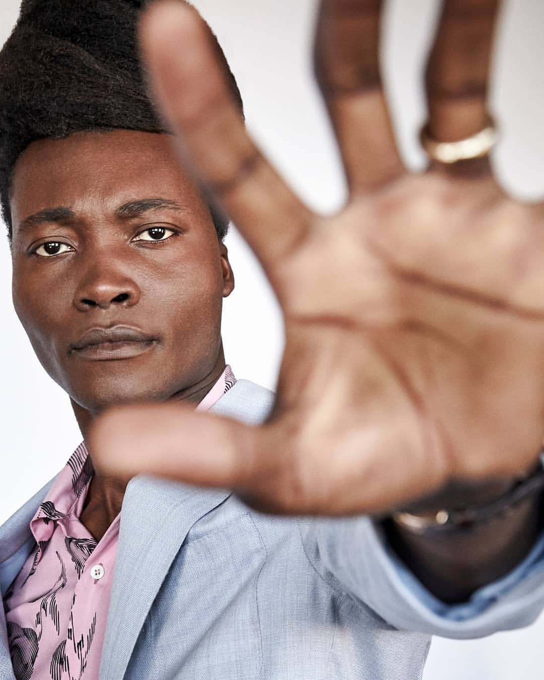 Portrait of an adult male with an afro hairstyle, wearing a blue blazer and floral shirt, reaching out with his hand towards the camera.