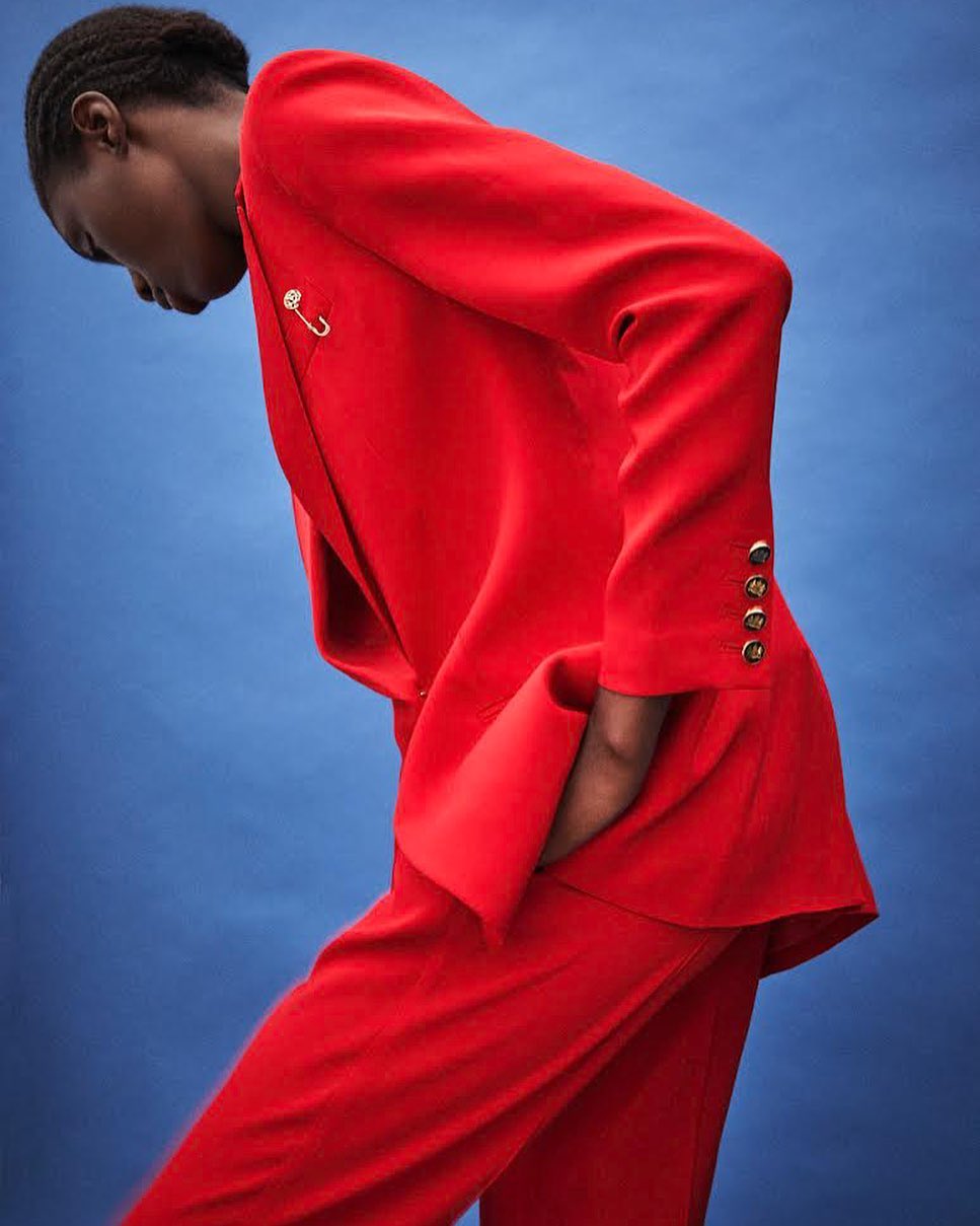 An African American woman wearing a red blazer and pantsuit poses against a solid blue backdrop in a fashion or beauty context.