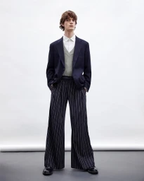 Full-length portrait of a male model wearing a navy blazer, patterned sweater vest, white shirt, and wide-legged pinstripe trousers against a grey seamless backdrop.