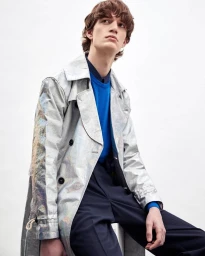 Young male model wearing a metallic trench coat and blue sweater against a plain white background