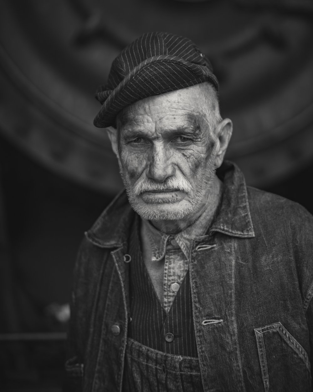 Monochrome portrait of an elderly man wearing a cap and overalls, with a weathered face and mustache, set against a dark background.