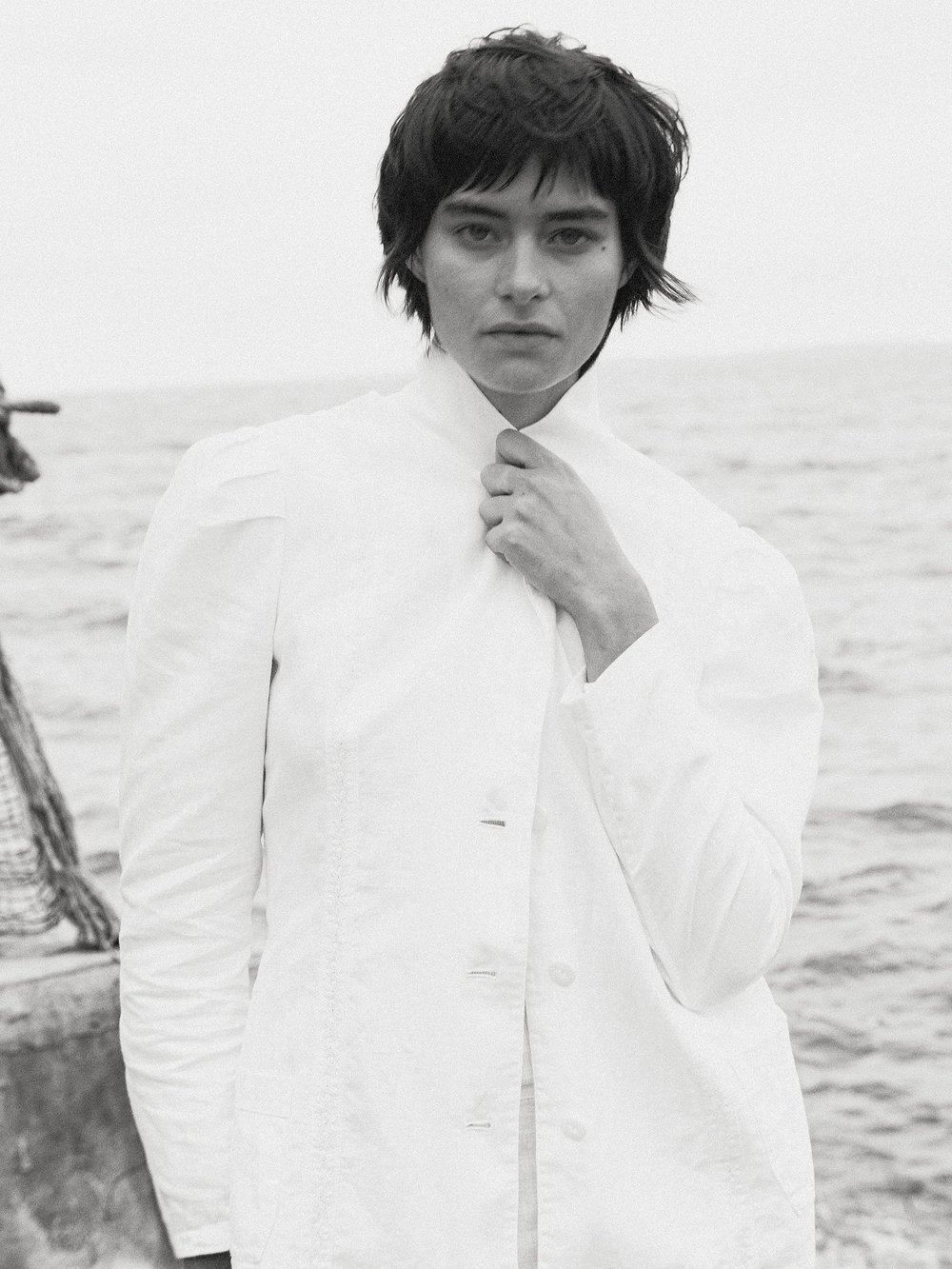Monochrome portrait of a fair-skinned adult woman in a white jacket against a blurred ocean backdrop, holding her jacket collar.