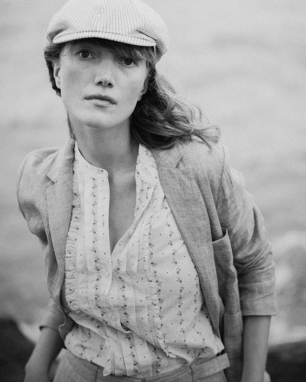 Monochrome portrait of a female model in a newsboy cap and linen blazer, posing outdoors with natural light and a serene expression.