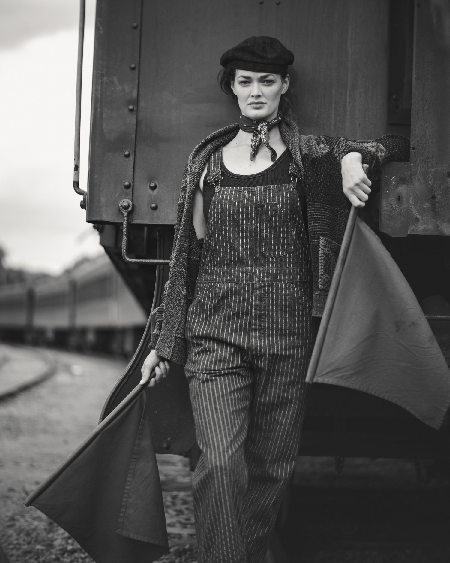 Black and white portrait of a female model wearing striped overalls, leaning against a vintage train car, holding red signal flags.