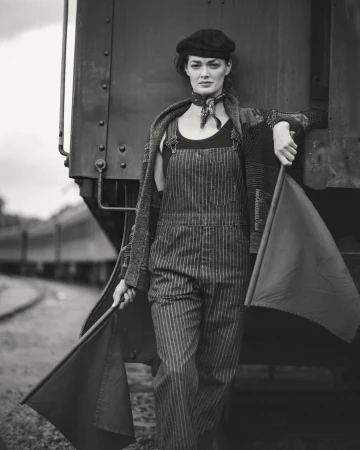 Black and white portrait of a female model wearing striped overalls, leaning against a vintage train car, holding red signal flags.