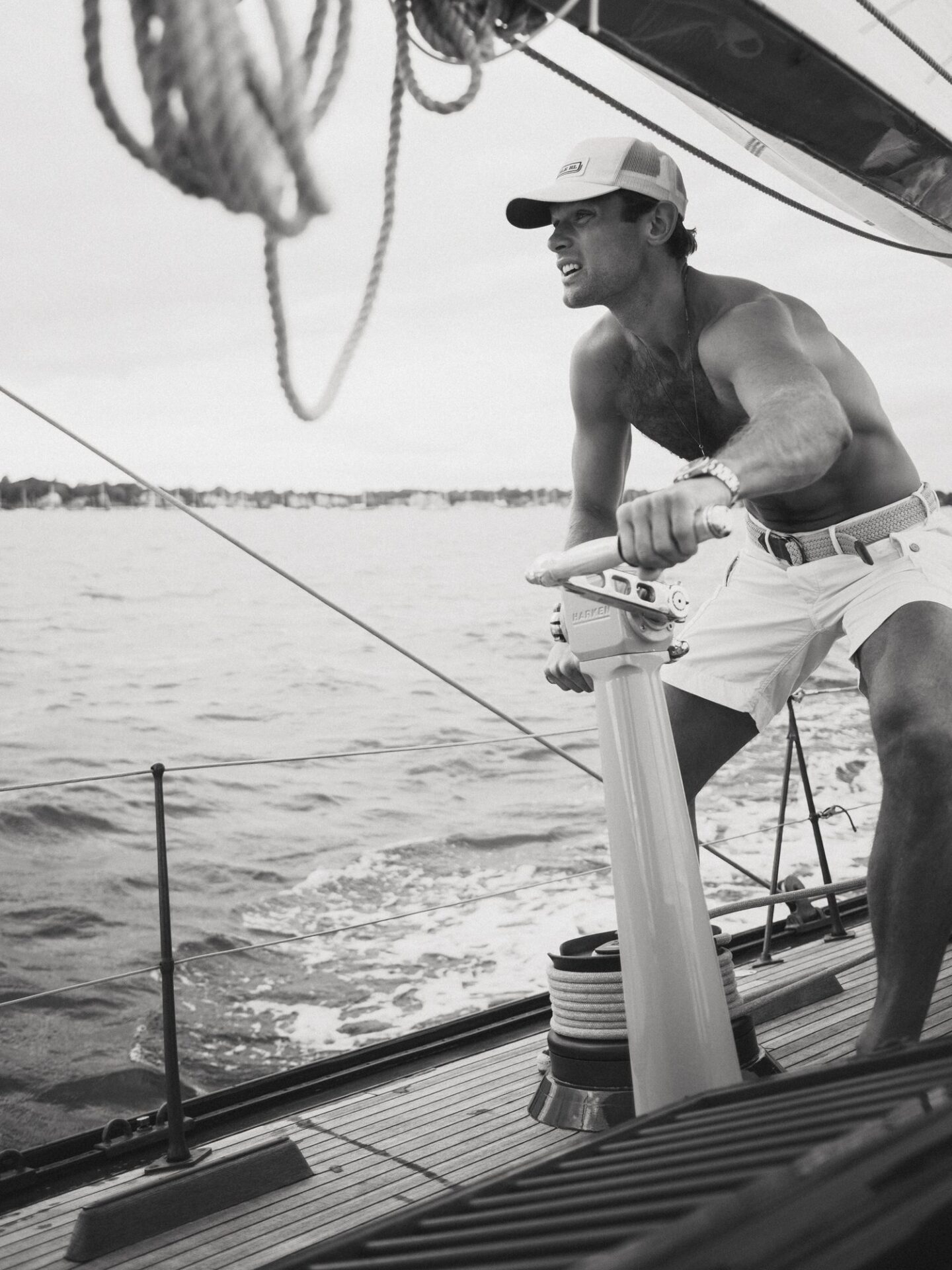 Monochrome shot of a man steering a sailboat, wearing shorts and a cap, with water and sky in the background, capturing a nautical scene.